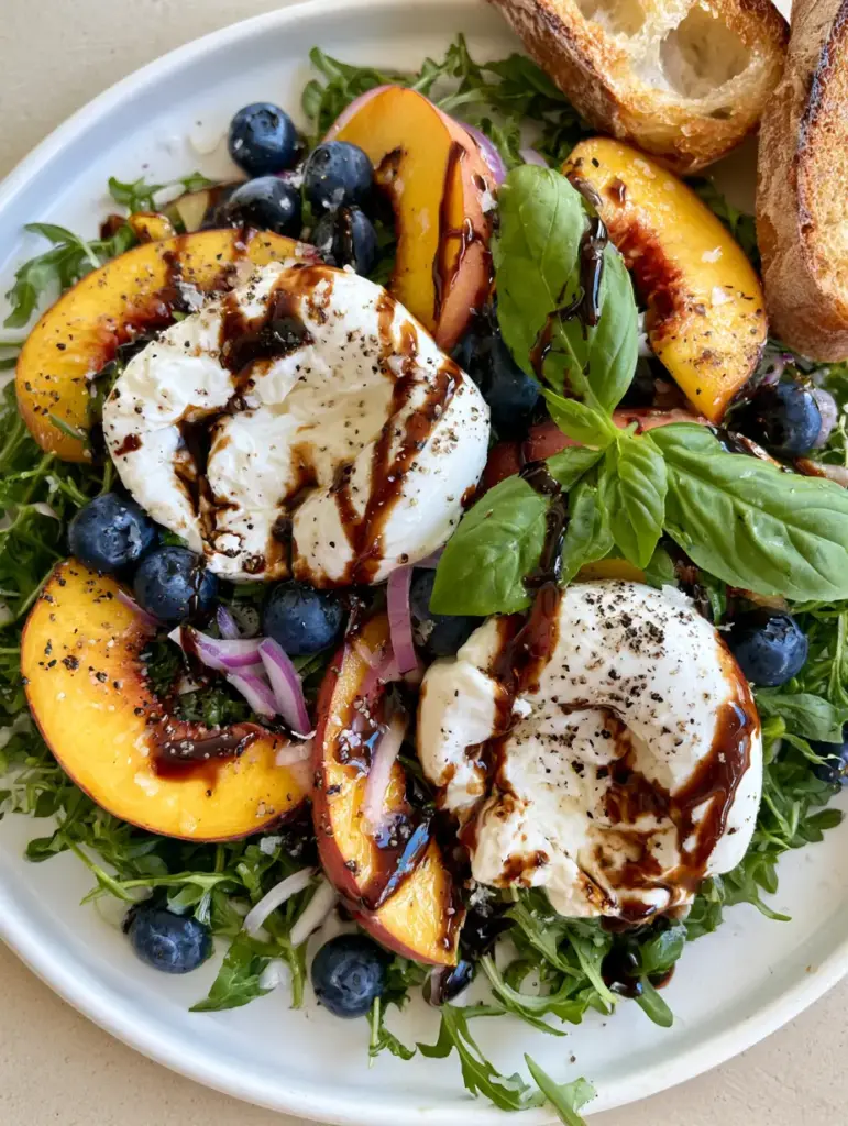 Peach Burrata Salad with Balsamic Glaze 1 Detailed view of torn burrata cheese revealing creamy stracciatella center over juicy yellow peaches and dark green arugula.
